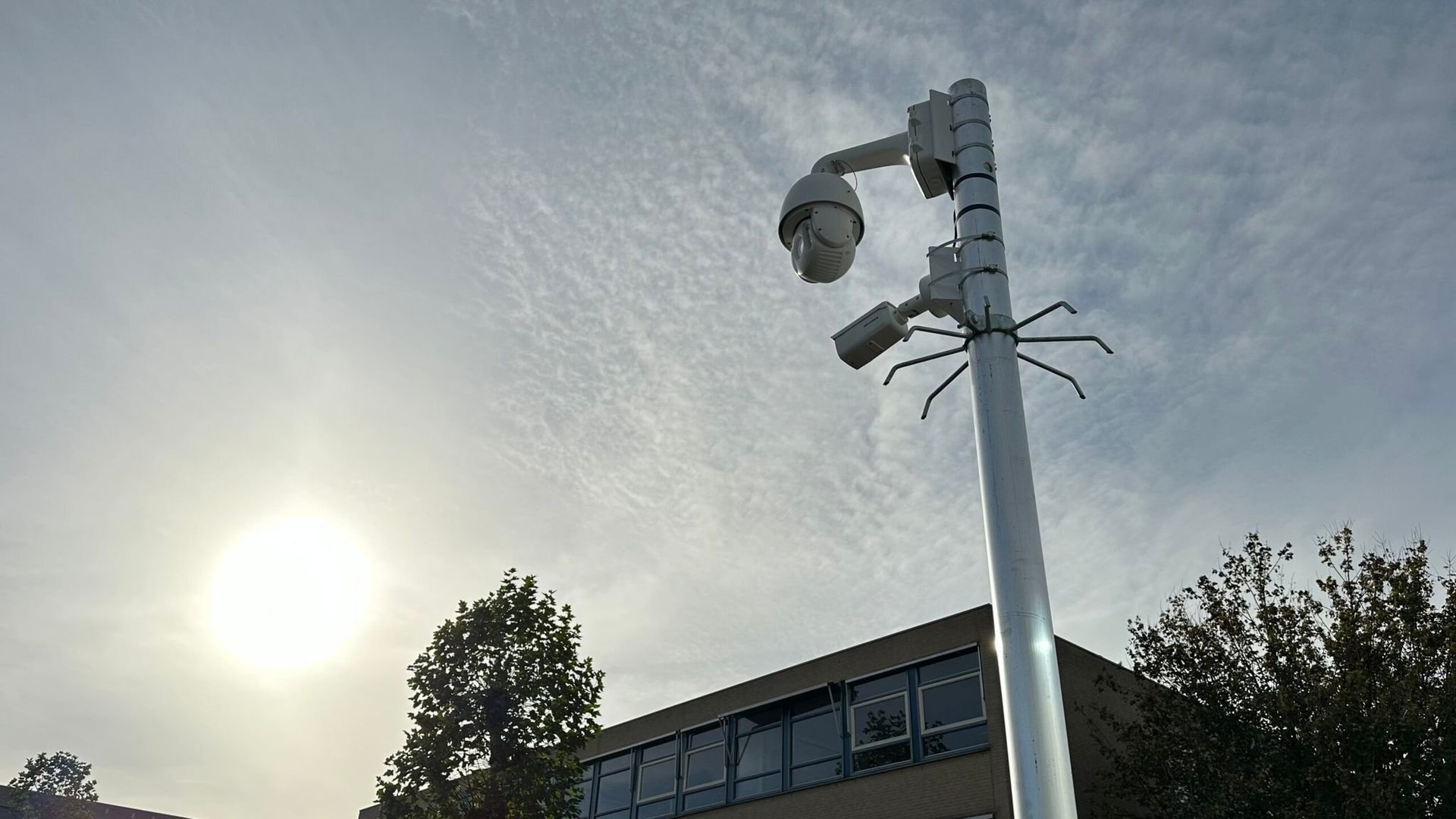 Surveillance camera mounted on tall metal pole against cloudy sky with building and trees below