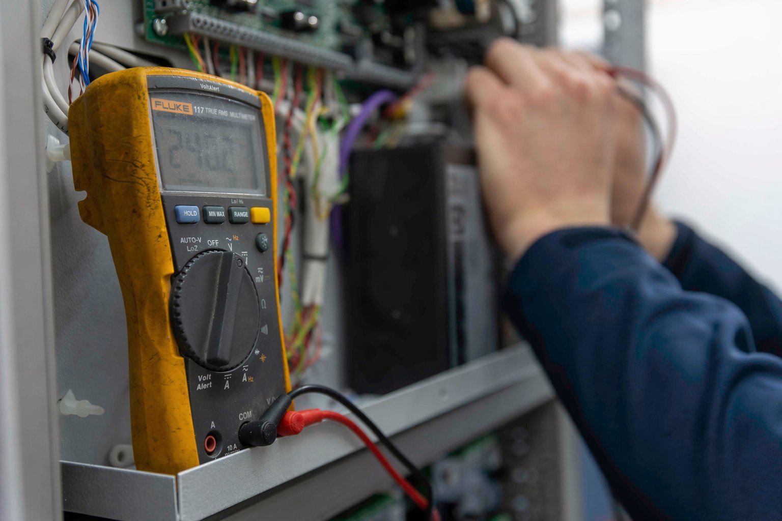 Technician using a yellow digital multimeter to test electrical connections on circuit board equipment